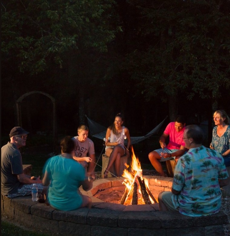 A group of seven adults are gathered around a stone firepit at night with roasting sticks in the fire